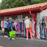 l'extérieur de la recyclerie SELgarrekin. On y voit des vêtements et des chaussures installés sur des portants, des personnes qui les regardent et le bâtiment de la recyclerie en arrière plan.